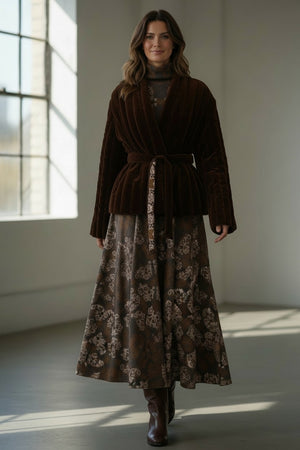 A woman stands indoors by a window, smiling softly with wavy hair. She wears the FRANCO FERRARI - REVERSIBLE KIMONO JACKET, which features a patterned velvet design and a subtle kimono silhouette, paired with a black top, black pants, and black loafers.