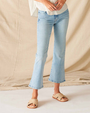 A person wears MERSEA’s Murphy Mini Boot Cut Jean in Sea Spray—light blue, 4-way stretch denim—paired with a white top and beige slide sandals, standing on a white floor before a beige fabric backdrop. Their face is not visible.