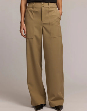A person stands facing away, wearing HINSON WU's KELLY TRAVELER'S TWILL PANT—high-waisted, wide-leg tan trousers with large back pockets—paired with a black top against a light neutral background.