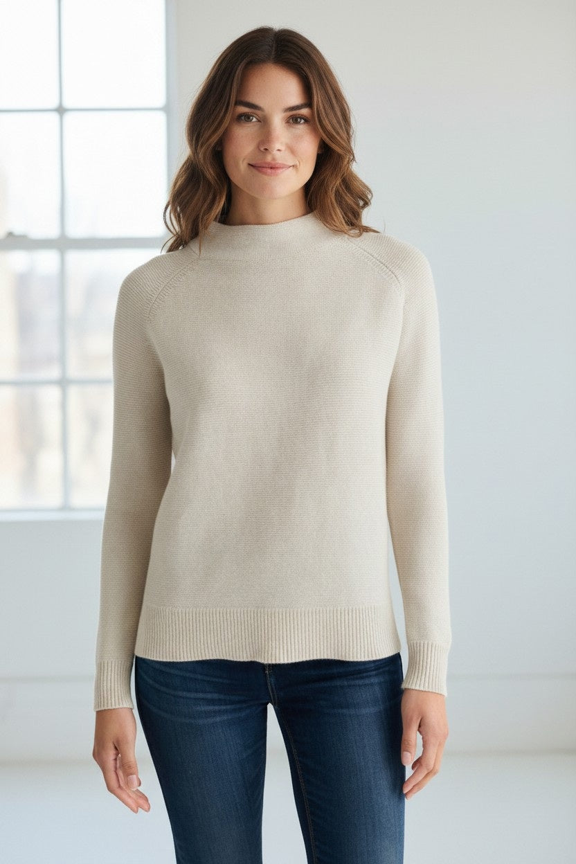 A woman with wavy brown hair, dressed in blue jeans and the KINROSS Garter Funnel Cotton Sweater in Cream by KINROSS, stands indoors in front of a window, offering a subtle smile to the camera. The classic-fit sweater features a mock neckline, blending style and comfort for her relaxed look.