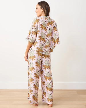 A woman sits on a white cube, wearing the PRINTFRESH - SATIN BAGHEERA WILDEST DREAM PAJAMA SET with leopard print. Her long hair cascades past a warm smile as she rests her left hand on her knee, bare feet gently placed on the light wood floor against a plain white wall.