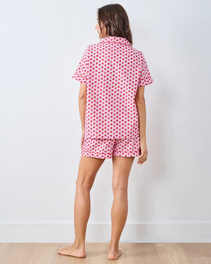 A person wearing the PRINTFRESH - MINI CANDY HEARTS PAJAMA SHORT SET by PRINTFRESH, featuring pink fabric with red heart patterns, stands with one hand in their shorts pocket. The image highlights the details of the pajama set and does not show the person's face.