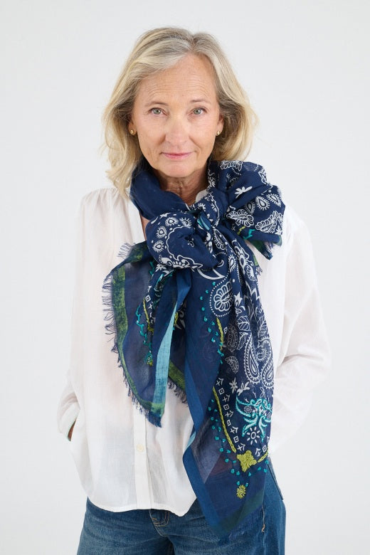A person with long gray hair displays the STORIATIPIC - KIKI SCARF IN MARINE by STORIATIPIC, a large sheer blue linen scarf with white floral and paisley patterns and delicate handmade embroidery, against a plain white background.
