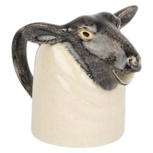 The QUAIL CERAMICS - BLACK SUFFOLK SHEEP JUG by QUAIL DESIGNS, shaped like a sheep's head and upper body with a handle at the back, sits on wood—ideal for farmhouse kitchens against a blurred green backdrop.