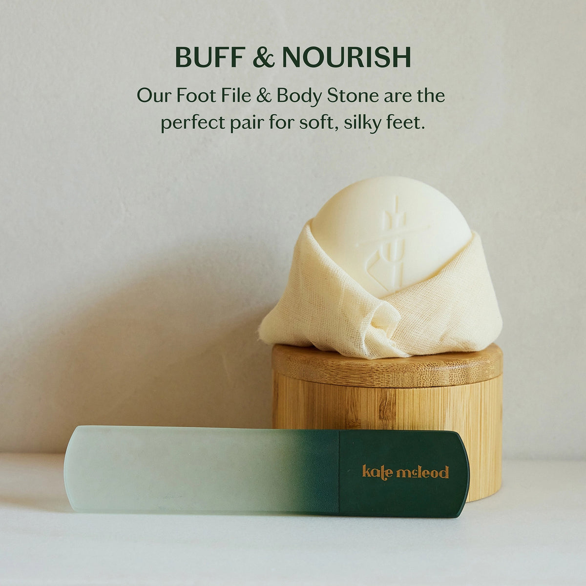 A hand with pale pink nails holds a cylindrical, green-gradient object labeled "KATE MCLEOD," designed for callus care—the light blue-green background enhances its minimalist aesthetic.