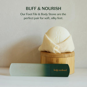 A hand with pale pink nails holds a cylindrical, green-gradient object labeled "KATE MCLEOD," designed for callus care—the light blue-green background enhances its minimalist aesthetic.