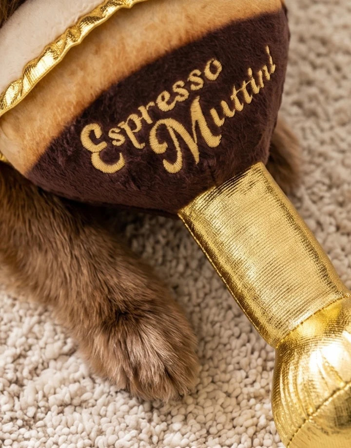 An espresso martini-shaped plush from HAUTE DIGGITY DOG, the ESPRESSO MUTTINI DOG TOY, rests on a marble table beside a coffee cup. This durable dog toy features gold, brown, and cream colors and displays “Espresso Mutini” on the side.