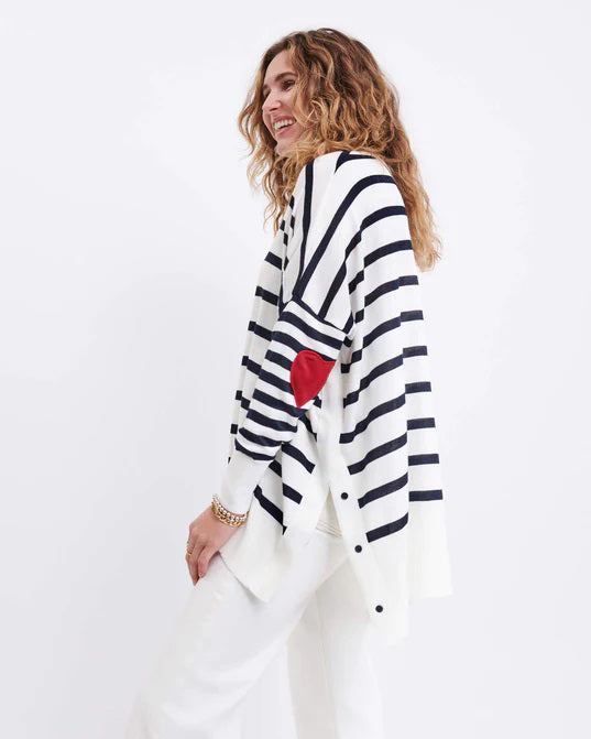 A woman with wavy blonde hair wears the MERSEA - AMOUR HEART SWEATER by MERSEA, a loose French-striped top with a red heart elbow patch. She pairs it with white pants and gold earrings, standing before a plain white background.