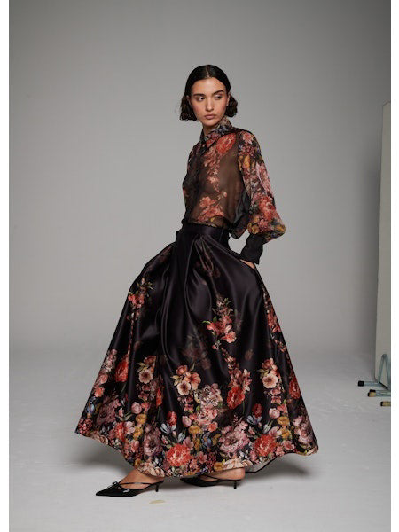 A woman poses in the MILVA MI - LONG FLORAL MAXI SKIRT by MILVA, paired with a sheer, long-sleeved floral blouse and pointed black heels against a simple studio backdrop.
