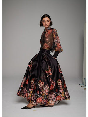 A woman poses in the MILVA MI - LONG FLORAL MAXI SKIRT by MILVA, paired with a sheer, long-sleeved floral blouse and pointed black heels against a simple studio backdrop.