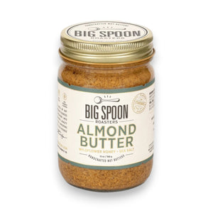 A glass jar of BIG SPOON ROASTERS - ALMOND BUTTER WILDFLOWER HONEY NUT BUTTER with a gold lid and a white label featuring branding and product details in black and gold text.