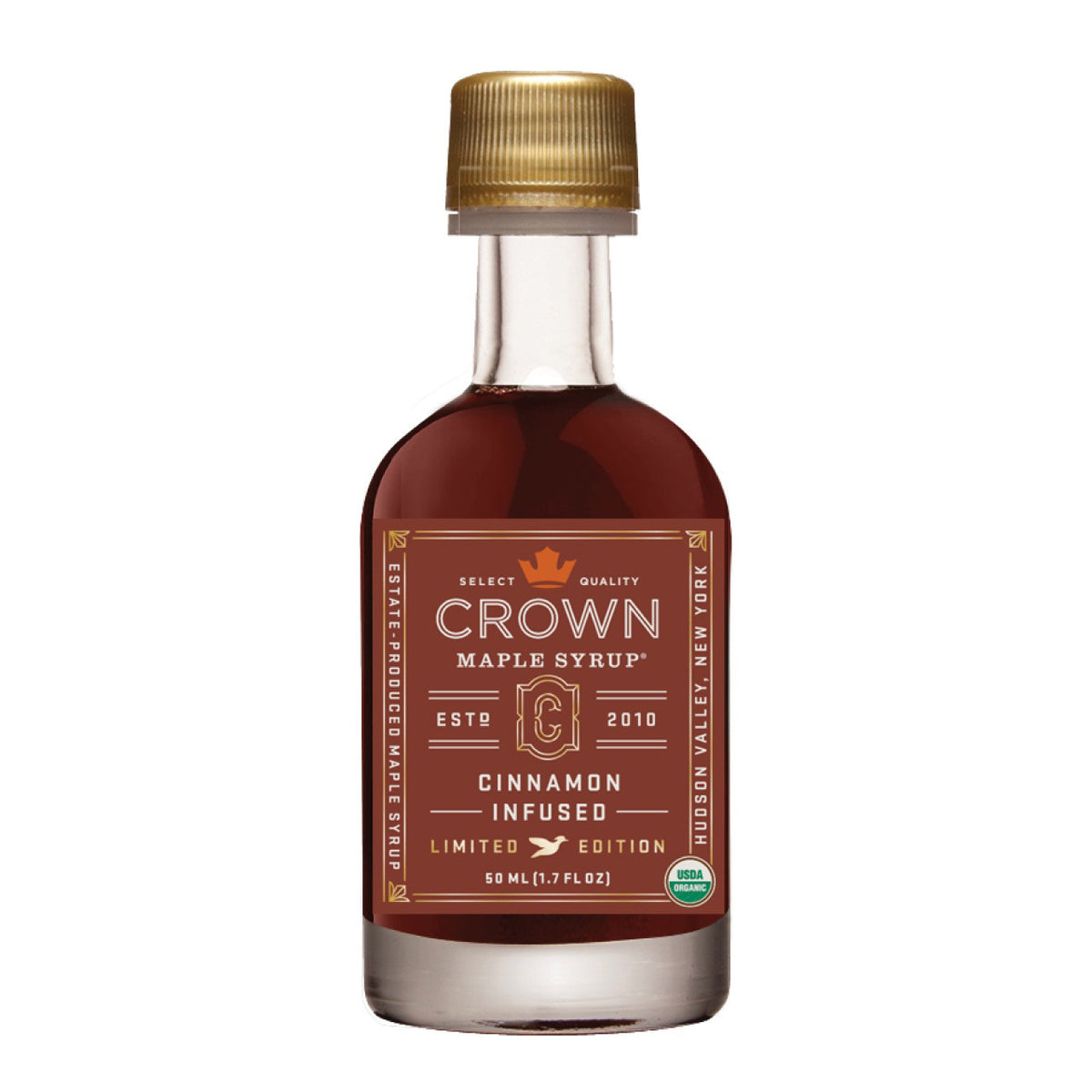 A limited edition small glass bottle of CROWN MAPLE - CINNAMON INFUSED MAPLE SYRUP. The bottle features a gold cap and a brown label with gold and white text detailing the organic maple syrup's status, its established year (2010), and its origin in Hudson Valley, New York.
