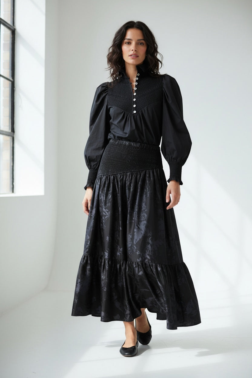 A woman with long brown hair faces away, wearing the HOPE FOR FLOWERS Smocked Shirt in Black with puffed sleeves and paired with a dark floral skirt, standing before a gray backdrop.