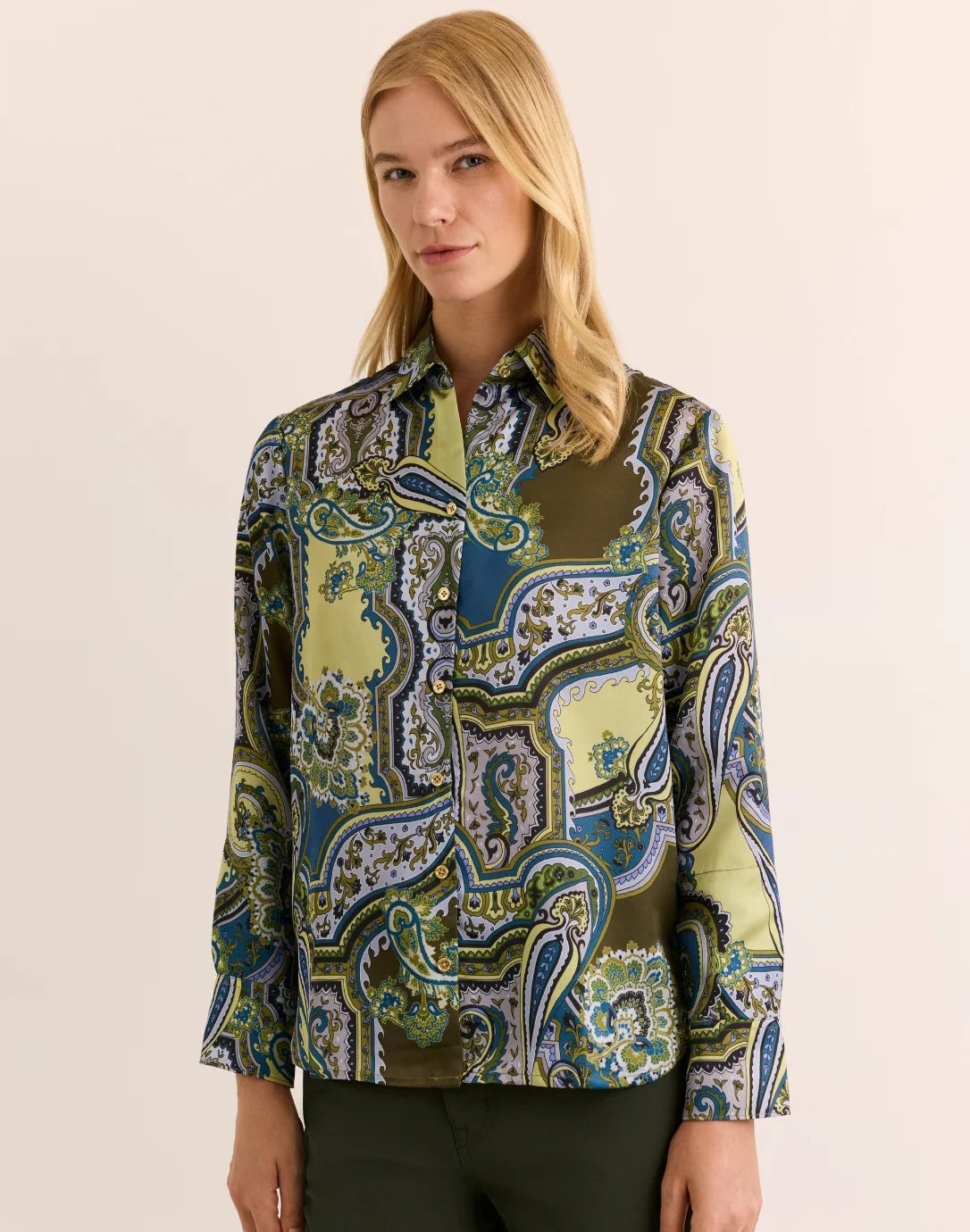 A woman with blonde hair wears the HINSON WU Margot Long Sleeve Paisley Shirt, a colorful button-up featuring green, blue, and yellow paisley patterns and crafted from luxurious Italian polyester for an elegant drape and silky feel. She pairs it with dark green pants and stands against a neutral background.