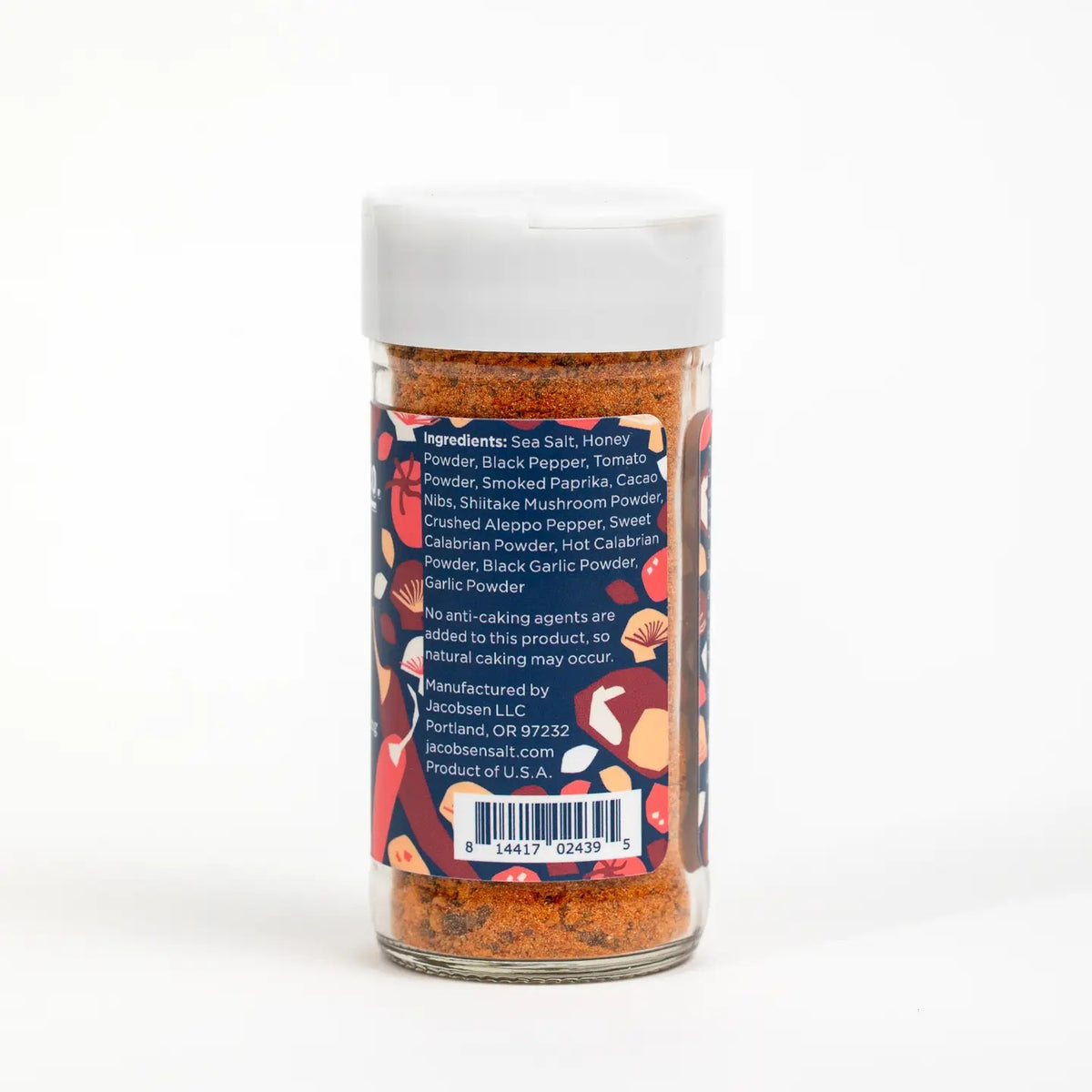A jar of JACOBSEN SALT CO. HOUSE SPECIAL SEASONING sits on a kitchen counter, surrounded by a ceramic bowl, a glass jar of honey, a bottle of oil, and wooden utensils.