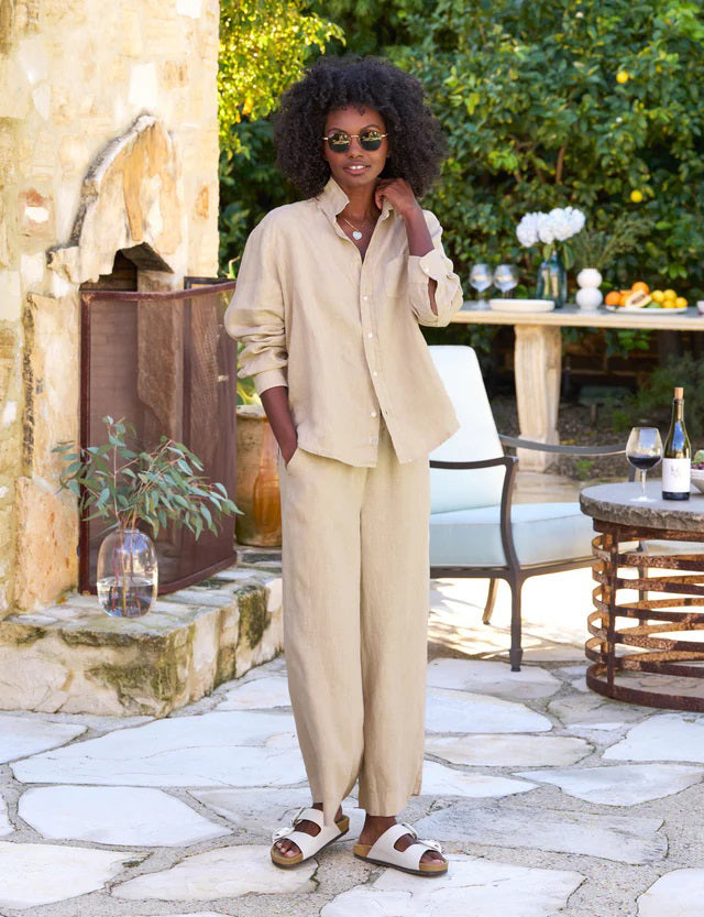 A woman with curly hair, wearing sunglasses, a beige long-sleeve shirt, and a matching pair of FRANK &amp; EILEEN Maisie Wide Leg Linen Pull On Pants from the Tuscany Getaway Set, stands outdoors. She is smiling and leaning against a rustic stone wall. Nearby is a table with a wine bottle and glass, and a white chair under a lush green tree.