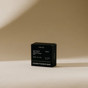 A white conditioner bar rests next to its black packaging on a beige background. The packaging displays "PRESTON," "Softening Argan Oil Conditioner Bar with Indian Amla Oil," "Amber," and "Made in USA," along with the weight "2.5 OZ" and "71 G." This plastic-free PRESTON - CONDITIONER BAR nourishes hair for a softer feel.
