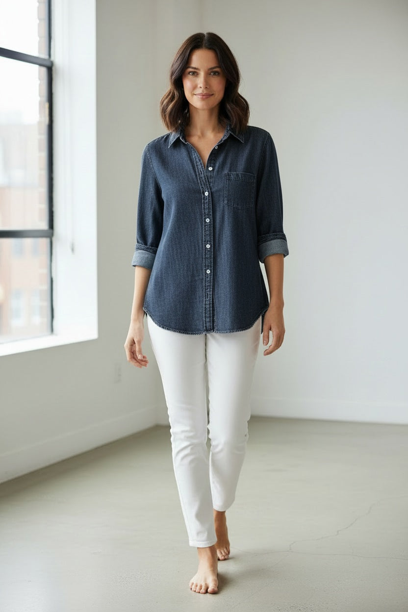 A woman with shoulder-length brown hair walks barefoot indoors, wearing the FRANK & EILEEN - EILEEN Relaxed Button Up Shirt in Famous Denim Washed Navy paired with white pants. She is in a bright, minimal room with large windows.