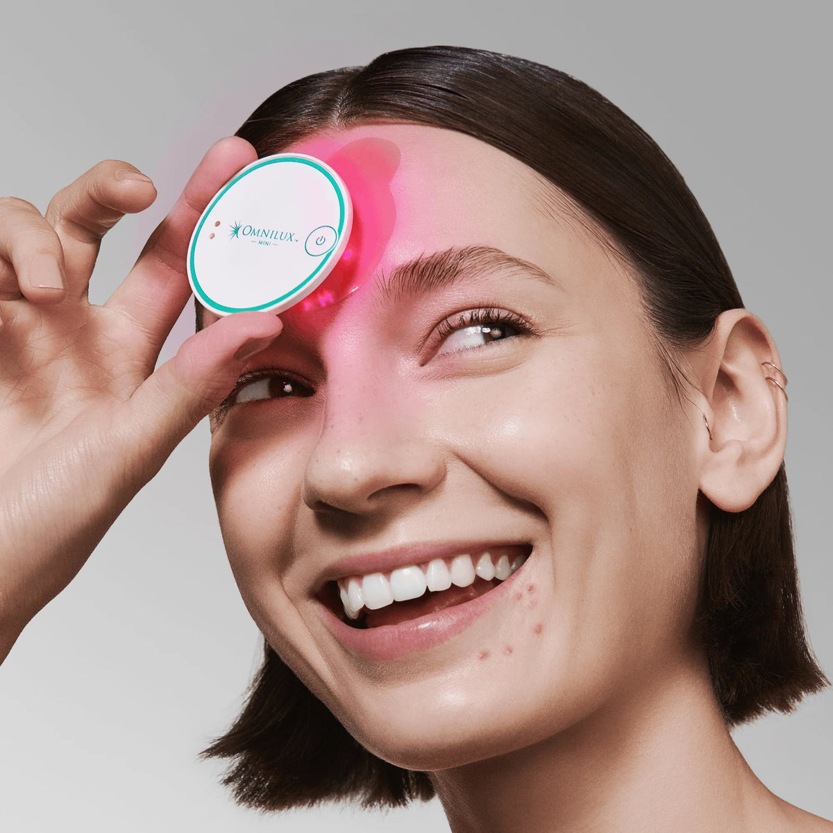 A round, white OMNILUX - BLEMISH ERASER KIT device from OMNILUX features a teal rim, power button, and logo; a soft pink light glows from underneath, showcasing its advanced blue and red light therapy technology.
