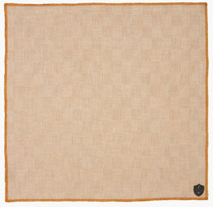 The CLIFTON WILSON - TAN CHECK LINEN POCKET SQUARE features a subtle check pattern, orange edging, and a small black shield emblem in one corner.