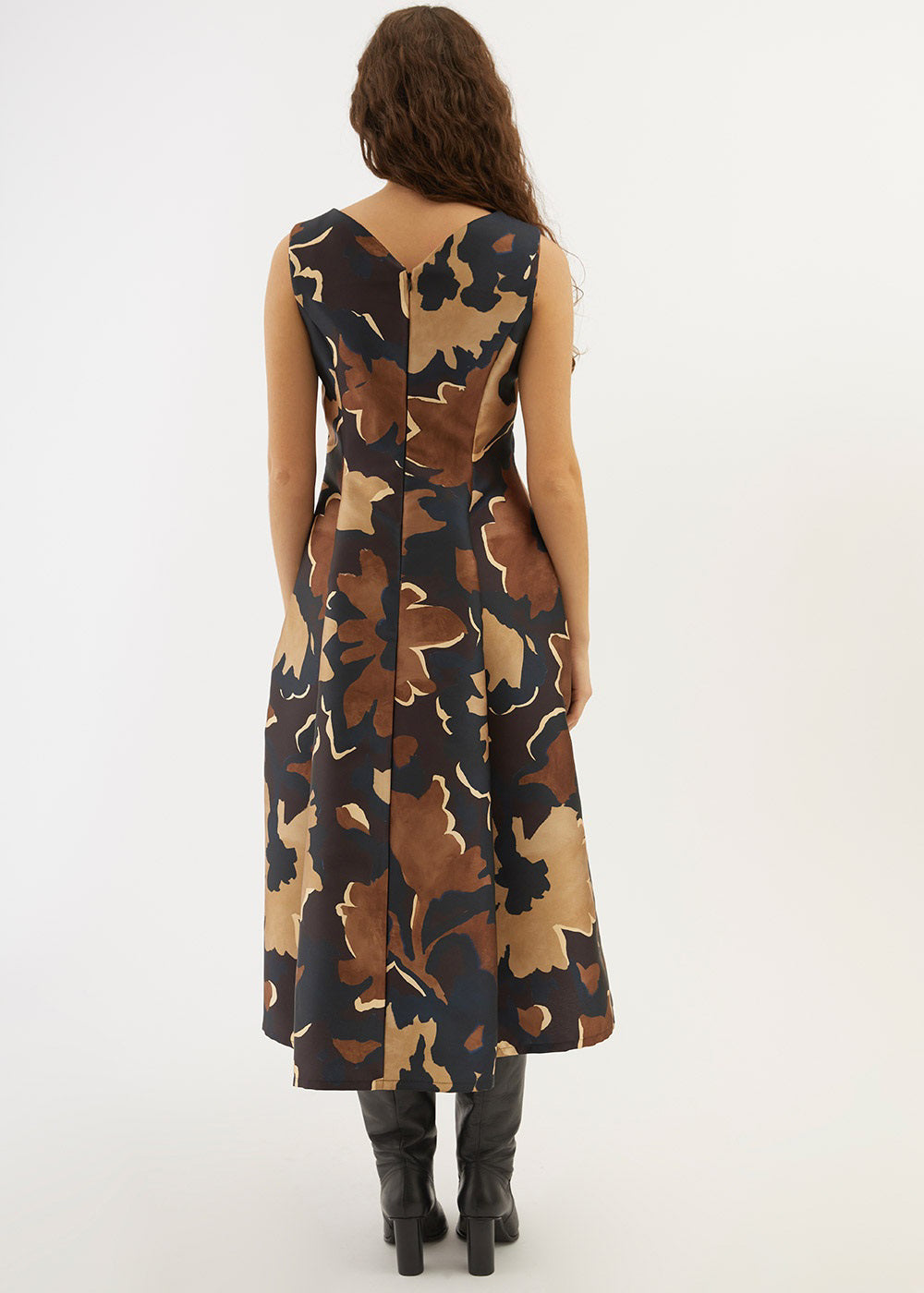 A woman stands with her hands in the pockets of the PARVATI DRESS by ALYKI LABORATORIO, a sleeveless, V-neck, calf-length dress featuring a brown and black abstract floral pattern, styled with black knee-high boots.