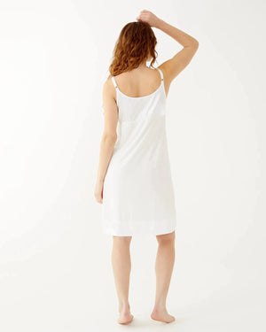 A woman with long wavy hair wears the sleeveless, knee-length MERSEA - MOON DANCER PAJAMA DRESS and smiles gently at the camera, barefoot against a plain white background—a perfect bridal gift.