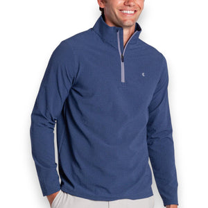 A man in the CRIQUET Seersucker Performance Pullover in Navy and light pants smiles with his hand by his side; this lightweight layer from CRIQUET offers UPF 50 protection against a simple background.