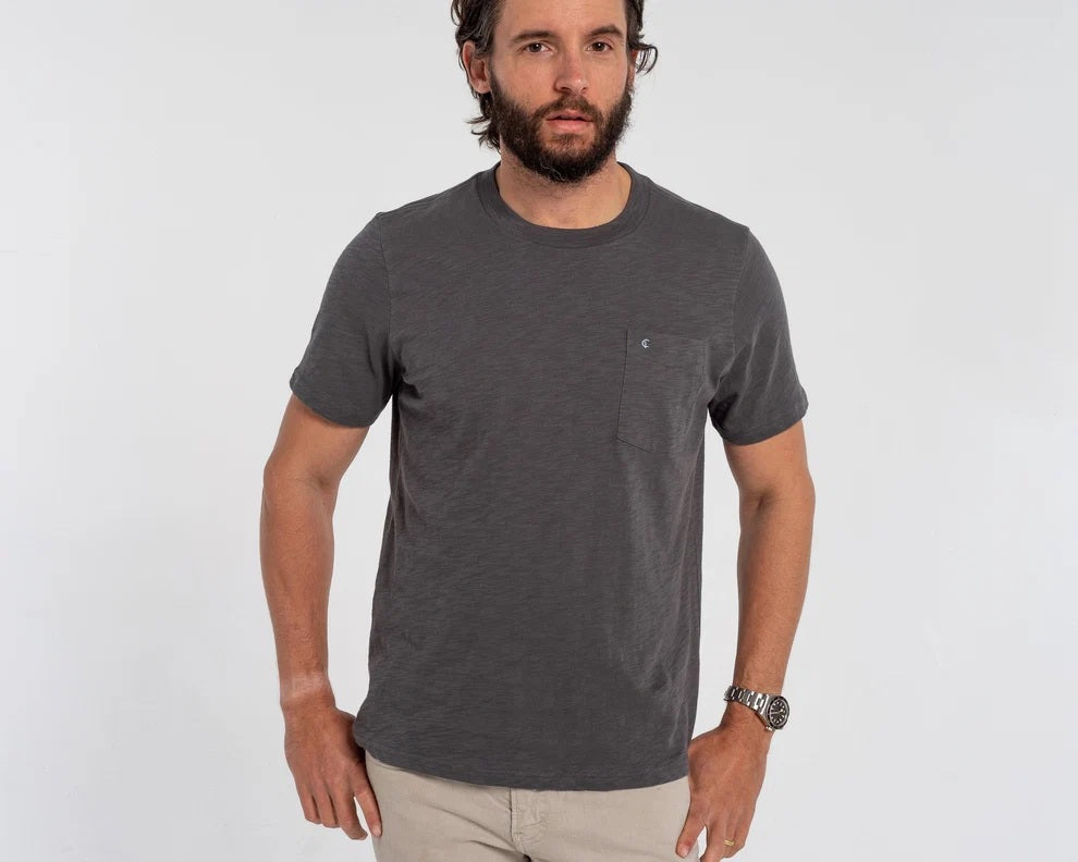 A man with a gray beard wearing the CRIQUET - SLUB COTTON SHORT SLEEVE POCKET T-SHIRT IN WASHED BLACK holds a canned drink in a green koozie outdoors, with trees and a white vehicle blurred in the background.