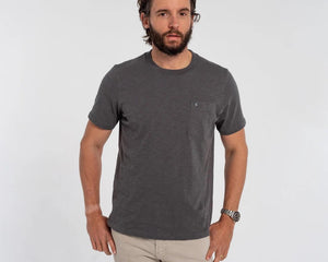 A man with a gray beard wearing the CRIQUET - SLUB COTTON SHORT SLEEVE POCKET T-SHIRT IN WASHED BLACK holds a canned drink in a green koozie outdoors, with trees and a white vehicle blurred in the background.