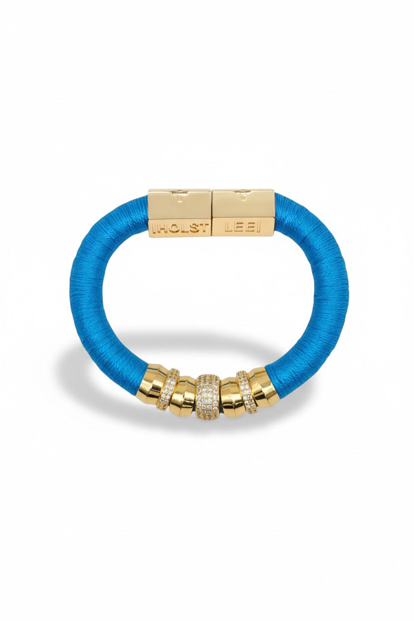 The HOLST + LEE - LUXE CLASSIC BRACELET by HOLST & LEE features an elegant orange cord with gold-plated accents, including a rectangular gold clasp adorned with several gold and diamond-encrusted beads near the clasp.