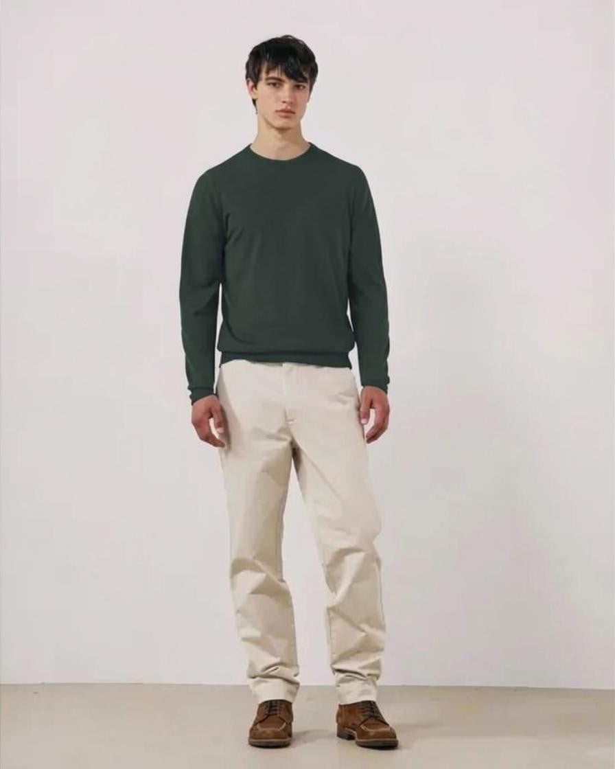 A young man stands with his hands in his pockets against a white background, wearing the HARTFORD Extra-Fine Merino Wool Crew Pullover in Forest and light-colored pants.