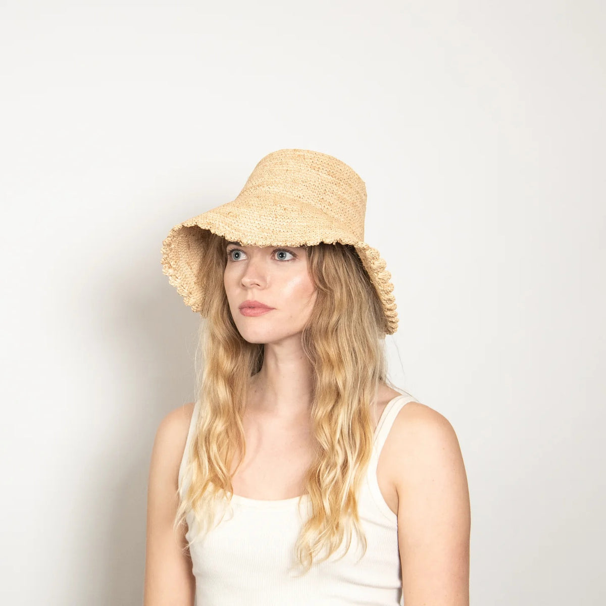 The WYETH Lacey Raffia Bucket Hat in Natural features a wide brim, textured woven pattern, and slightly scalloped edge. Displayed against a plain white background, this stylish hat is crafted by WYETH.