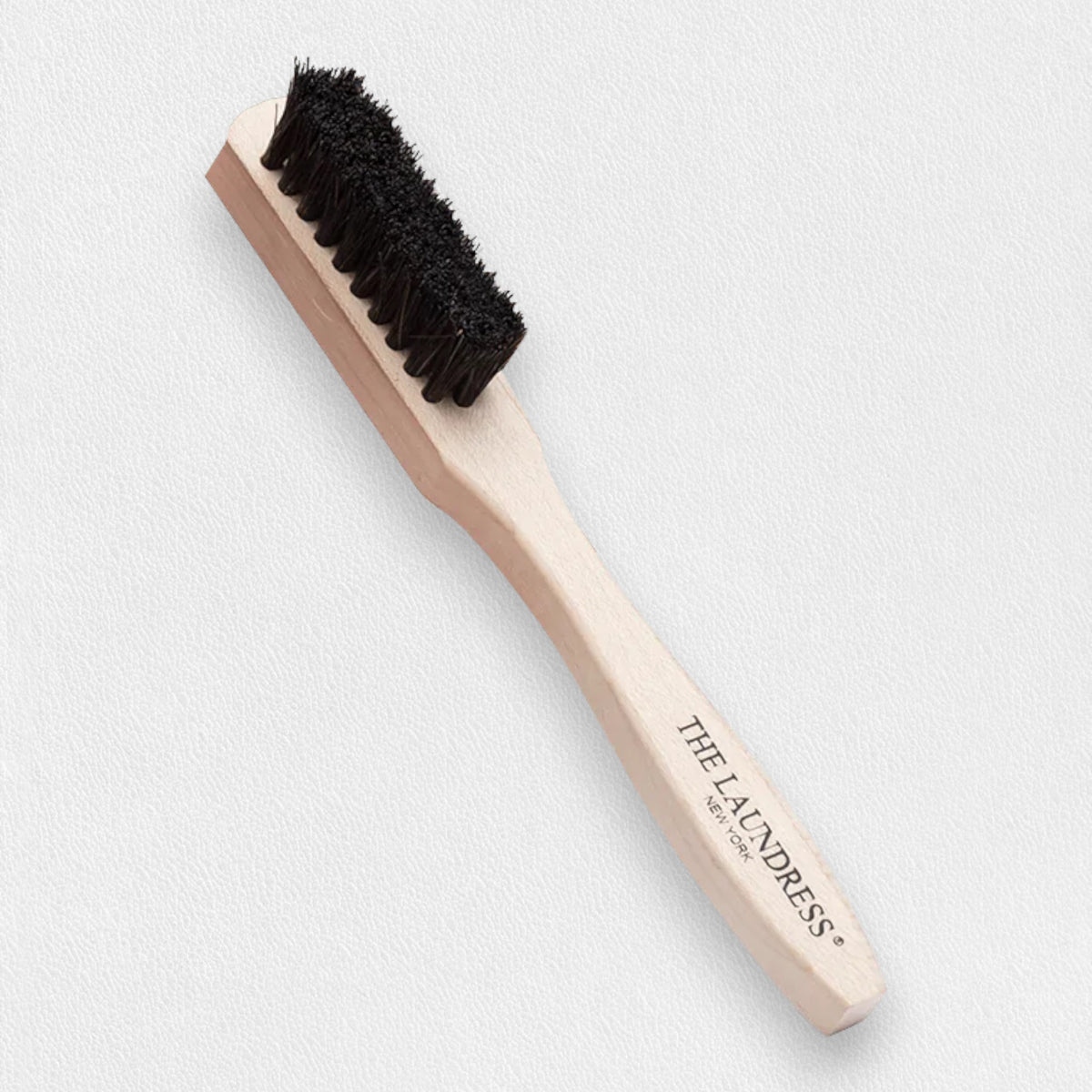 The Laundress - Stain Brush, a wooden brush with black bristles, is ideal for effective stain removal. Featuring a light-colored handle with &quot;The Laundress&quot; printed in black, this tool stands out against a plain, light-colored background, highlighting its importance in fabric care.