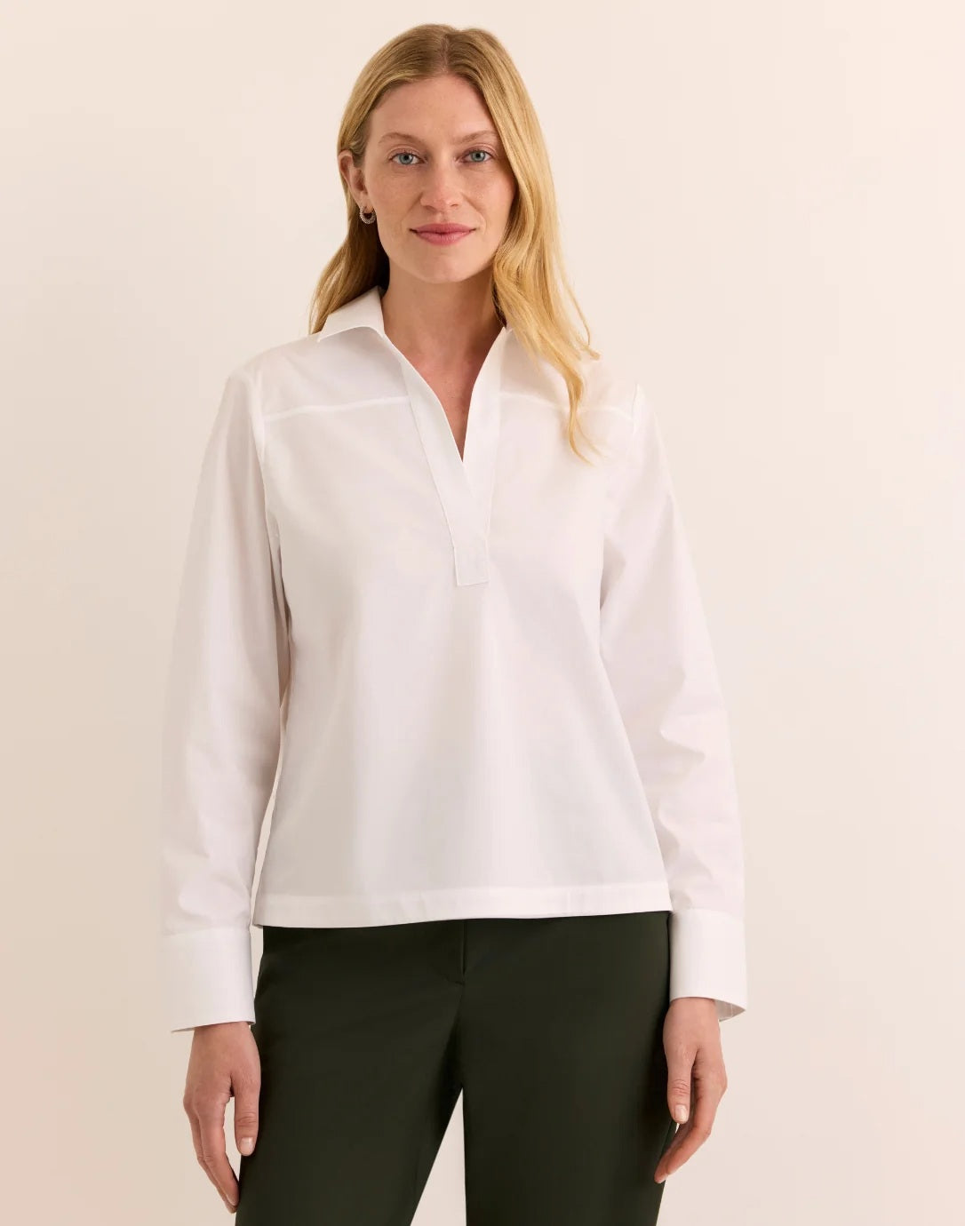 A woman with long blonde hair models the HINSON WU Monica Long Sleeve Button Back Top in white—a collared stretch polished cotton blouse featuring a split neckline and long sleeves—paired with dark green pants, standing against a plain light background.