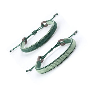 A close-up of a person's wrist wearing the MAKARLA MEN'S REVERSIBLE SURFER BRACELET in mint/hunter green. The person is dressed in a green shirt and blue jeans with a small tear near the pocket.