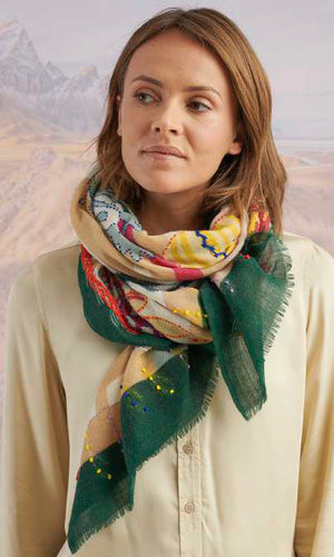 A person holds up the STORIATIPIC - ANDRES WOOL SCARF, an elegant rectangular accessory by STORIATIPIC, featuring colorful potted plants and vases with an orange border against a blurred natural backdrop.