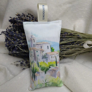 A L'AUGUSTE PROVENCE French Lavender Sachet from Provence, adorned with a cream "L’AUGUSTE" ribbon and painted with a hillside village, hangs before dried lavender tied with white ribbon.