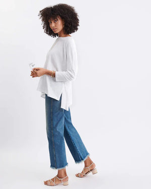 A woman with long, straight hair stands against a white wall, wearing the MERSEA Catalina Slub Tee in blue paired with white pants. The midweight textured cotton tee is from MERSEA. She looks at the camera with a neutral expression.