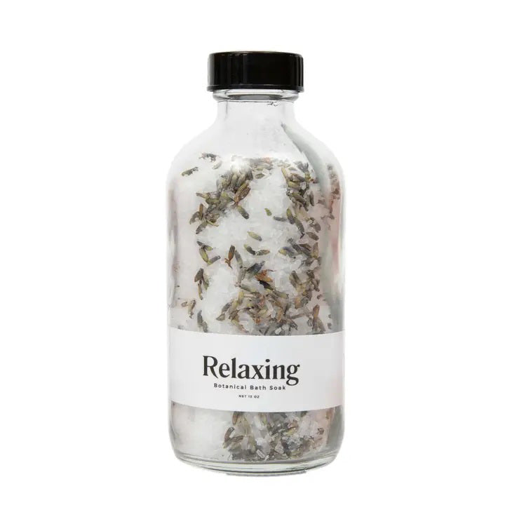A transparent glass bottle with a black cap contains a mixture of botanical bath soak ingredients, including dried lavender flowers and refreshing peppermint. The label on the bottle reads &quot;BOTANICAL BATH SOAK - RELAXING&quot; by GIFTEN MARKET.