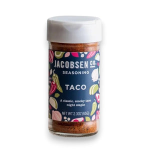 A jar of JACOBSEN SALT - TACO SEASONING from JACOBSEN SALT CO. rests on a kitchen shelf, alongside a white container, wooden utensils, a glass jar with a yellow lid, and a mason jar filled with honey.
