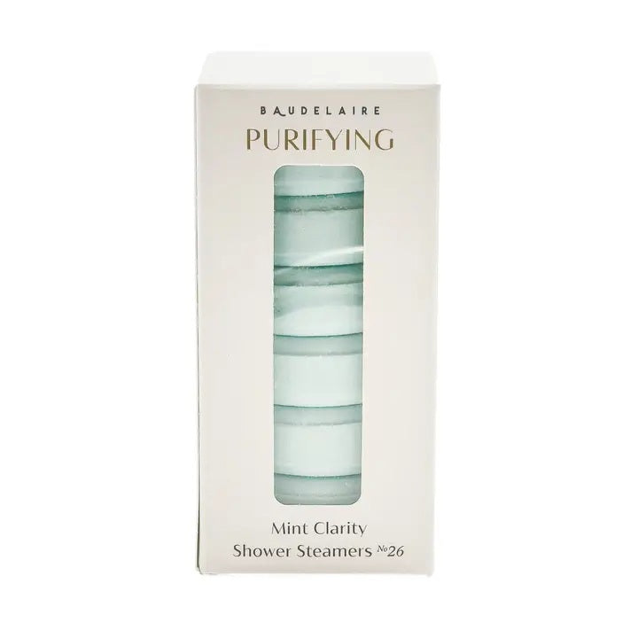 A white box labeled “BAUDELAIRE - PURIFY MINT CLARITY SHOWER STEAMER” contains light blue shower steamers stacked neatly inside, crafted for aromatherapy by BAUDELAIRE. The steamers, infused with essential oil blends, are visible through a clear window, and “Mint Clarity Shower Steamers No. 26” appears at the bottom of the box.