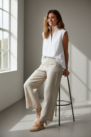 A woman in the FRANK & EILEEN OLIVIA Popover Henley Tank in white from FRANK & EILEEN sits on a stool by a sunlit window, smiling, wearing beige wide-leg pants, neutral-toned espadrille sandals, and her hair styled in relaxed, natural waves.