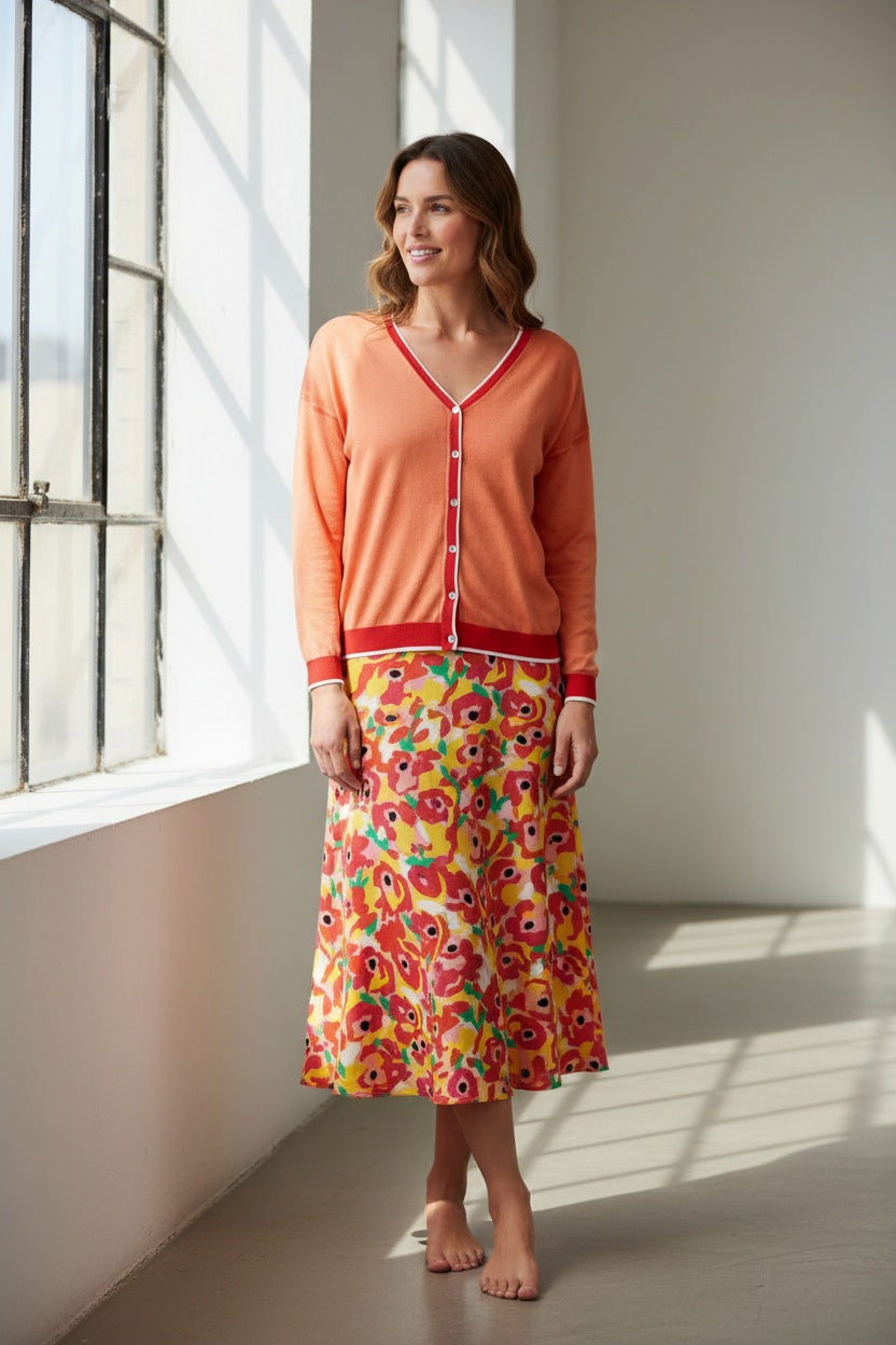 A woman wears the IN BED WITH YOU - BATWING SLEEVE CARDIGAN by IN BED WITH YOU/CLAUDIOLAI SRL, an orange V-neck with red and white trim, buttoned down the front, styled with multicolored patterned pants against a plain white background.