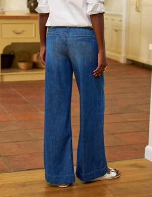 A person stands indoors on a tiled floor, wearing FRANK & EILEEN's West Village NYC Wide Leg Trouser in Italian Dream Light Denim 1986 Wash, paired with a white long-sleeve blouse and white sandals. The photo captures their lower half from behind.