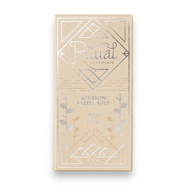 The image shows a light brown rectangular package of Ritual Chocolate labeled "RITUAL CHOCOLATE - BOURBON BARREL AGED CHOCOLATE BAR 75%." This limited edition chocolate, crafted from aged cacao nibs, features geometric designs in silver and white and contains 2.12 oz (60g) of chocolate. The background is a marble surface.