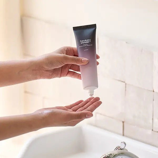 A person squeezes ASIAN BEAUTY WHOLESALE BLACK RICE TRIPLE AHA GENTLE CLEANSING GEL 100ML from a pink and black tube onto their hand over a sink, with a tiled wall in the background. The cleanser uses natural surfactants for gentle care.