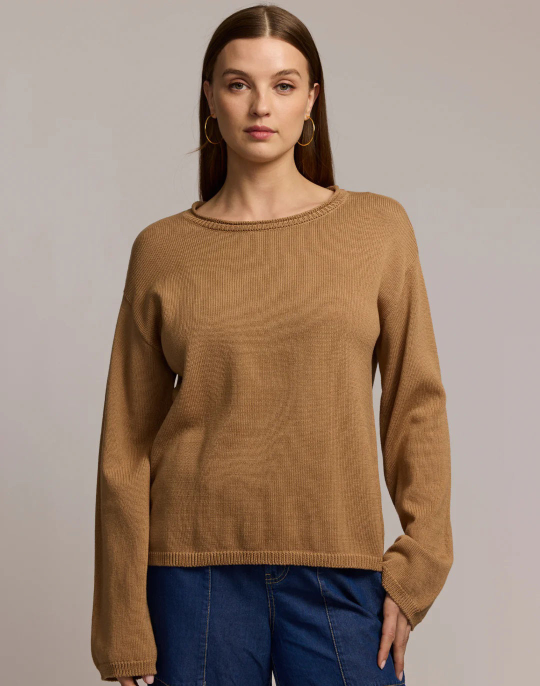 A woman with long brown hair and large hoop earrings models the HINSON WU AUDREY Long Sleeve Cotton Silk Sweater in tan with blue jeans, standing against a plain beige background, one hand raised near her shoulder.