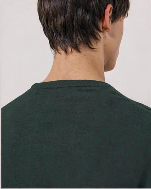 A young man stands with his hands in his pockets against a white background, wearing the HARTFORD Extra-Fine Merino Wool Crew Pullover in Forest and light-colored pants.
