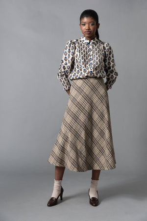 A woman stands in a dimly lit, ornately decorated room, wearing the HOPE FOR FLOWERS - CIRCLE SKIRT IN NATURAL CHECK by HOPE FOR FLOWERS. Warm light from a floor lamp casts shadows on the teal wall behind her.