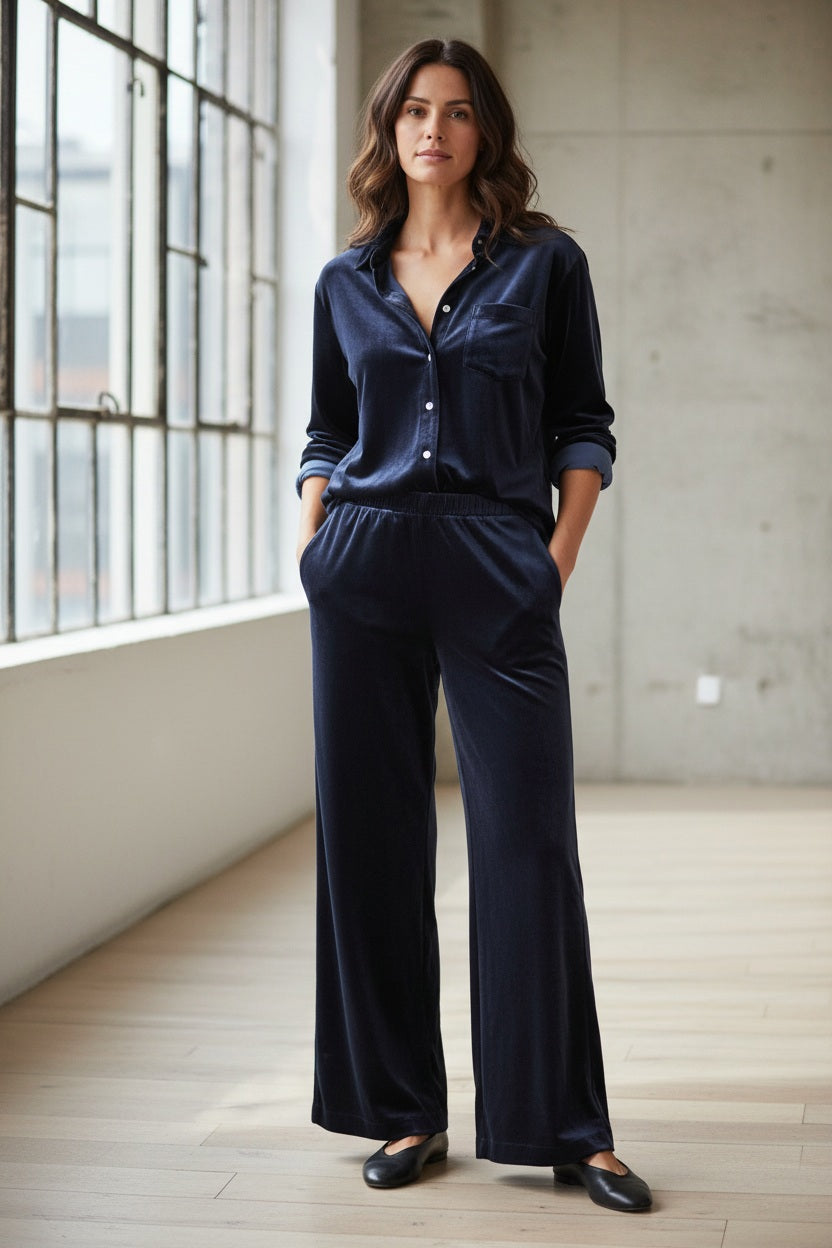 A woman with long wavy hair stands indoors by large windows, her hands in her pockets as she wears a navy blue button-up shirt paired with the FRANK & EILEEN MAISIE Velvet Wide Leg Pull On Pant in Classic Navy and black flats.